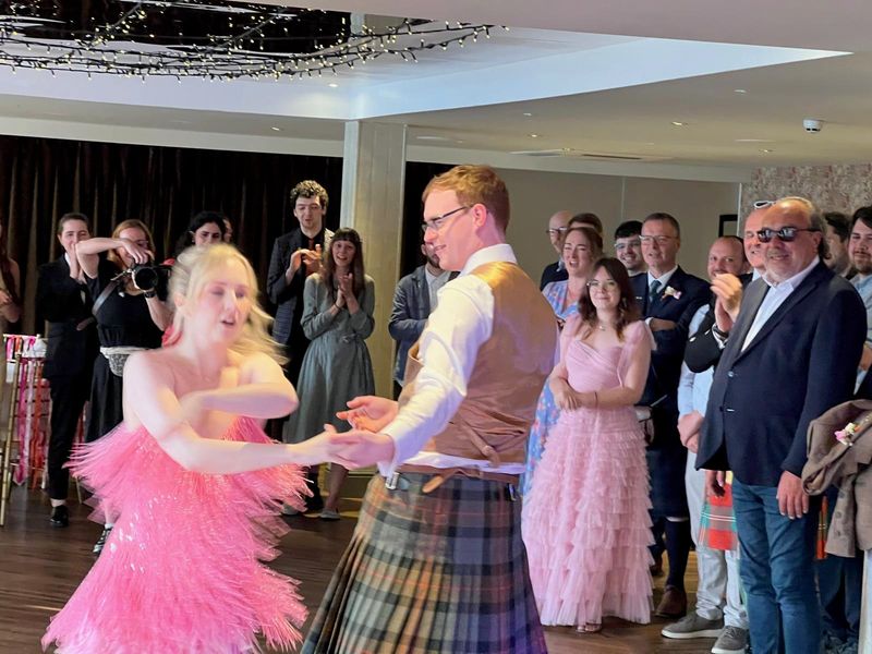 A guest in a pink gown spins with a kilted dancer mid-ceilidh